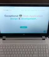 A laptop computer with a website displayed on the screen featuring the text 'Exceptional Web Application design & development'. The laptop is on a dark surface, and the website has a clean and modern design with a light background and colorful text elements.