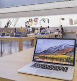 The image features a modern electronics store interior with wooden tables displaying various laptop models. Several people are present, some trying out devices while others converse. Large promotional images of smartwatches and smartphones are visible on the walls, adding to the store's sleek and innovative atmosphere.
