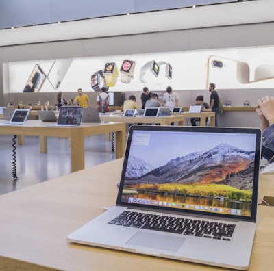 The image features a modern electronics store interior with wooden tables displaying various laptop models. Several people are present, some trying out devices while others converse. Large promotional images of smartwatches and smartphones are visible on the walls, adding to the store's sleek and innovative atmosphere.