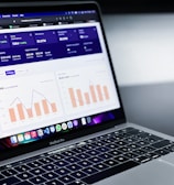 A laptop with an open screen displaying dashboard analytics featuring charts, graphs, and numerical data related to sales and subscriptions. The interface is predominantly dark-themed with sections highlighting key performance metrics. The device shown is a MacBook Pro, indicated by the keyboard layout and branding.