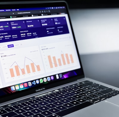 A laptop with an open screen displaying dashboard analytics featuring charts, graphs, and numerical data related to sales and subscriptions. The interface is predominantly dark-themed with sections highlighting key performance metrics. The device shown is a MacBook Pro, indicated by the keyboard layout and branding.