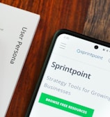 A wooden table with several documents and a smartphone. The documents are titled 'User Persona', 'Brand Colours', and 'Sprintpoint'. The smartphone displays a webpage for 'Sprintpoint', which offers strategy tools for growing businesses.