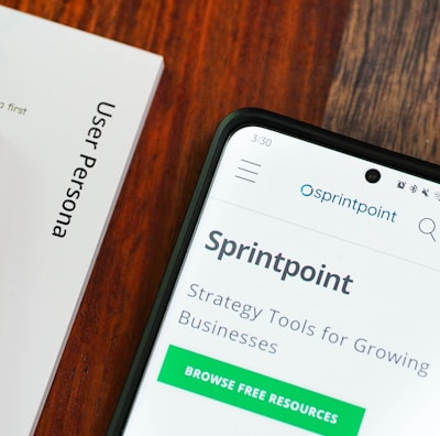 A wooden table with several documents and a smartphone. The documents are titled 'User Persona', 'Brand Colours', and 'Sprintpoint'. The smartphone displays a webpage for 'Sprintpoint', which offers strategy tools for growing businesses.