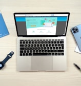 A laptop computer displaying a website on its screen is placed on a light wooden surface. Nearby are a blue smartphone, a smartwatch with a dark blue band, a blue folder, a white notepad, and a black pen.