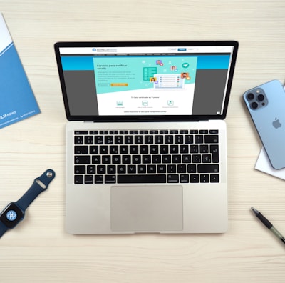 A laptop computer displaying a website on its screen is placed on a light wooden surface. Nearby are a blue smartphone, a smartwatch with a dark blue band, a blue folder, a white notepad, and a black pen.