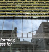 Glass-fronted building with reflections of the sky and nearby structures. Rows of greenery are integrated into the design above the windows. Large text on the building's facade reads 'Transforming Business for Good.'