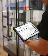 A person holds a tablet displaying a digital catalog in a retail store. The background features glass display cases with various products, and the store interior includes hardwood flooring.