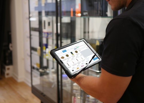 A person holds a tablet displaying a digital catalog in a retail store. The background features glass display cases with various products, and the store interior includes hardwood flooring.