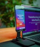 A smartphone is mounted on a stand, displaying a website with colorful graphics and the text 'Transforming Ideas into Reality'. Beside it, a laptop screen shows a blurred version of the same website. The background is soft and out of focus, with green foliage adding a hint of nature.