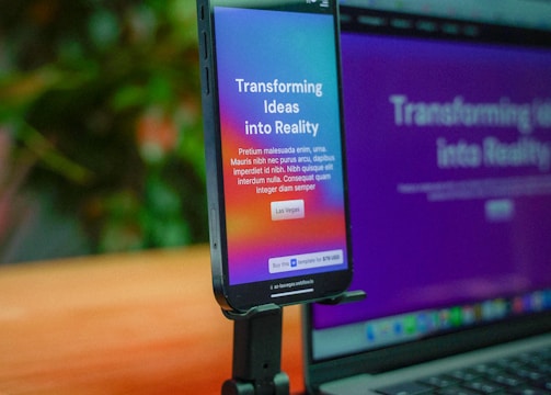 A smartphone is mounted on a stand, displaying a website with colorful graphics and the text 'Transforming Ideas into Reality'. Beside it, a laptop screen shows a blurred version of the same website. The background is soft and out of focus, with green foliage adding a hint of nature.